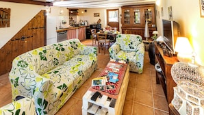 Un salon avec des canapés à motifs floraux, une table basse en bois sur laquelle sont posés des livres, un téléviseur et, au fond, un coin repas.