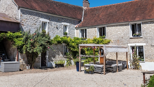 Un bâtiment en pierre avec un toit recouvert de tuiles rouges, une cour en gravier et une pergola en bois avec des sièges.