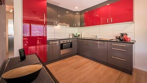 A modern kitchen with red and grey cabinetry, a built-in oven, and a dining area with a wooden table and chairs. - Apartment Breithorn Grindelwald (Grindelwald)