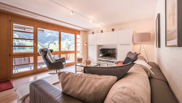 A modern living room with a large window, a sofa, a TV, and a lamp. - Apartment Breithorn Grindelwald (Grindelwald)