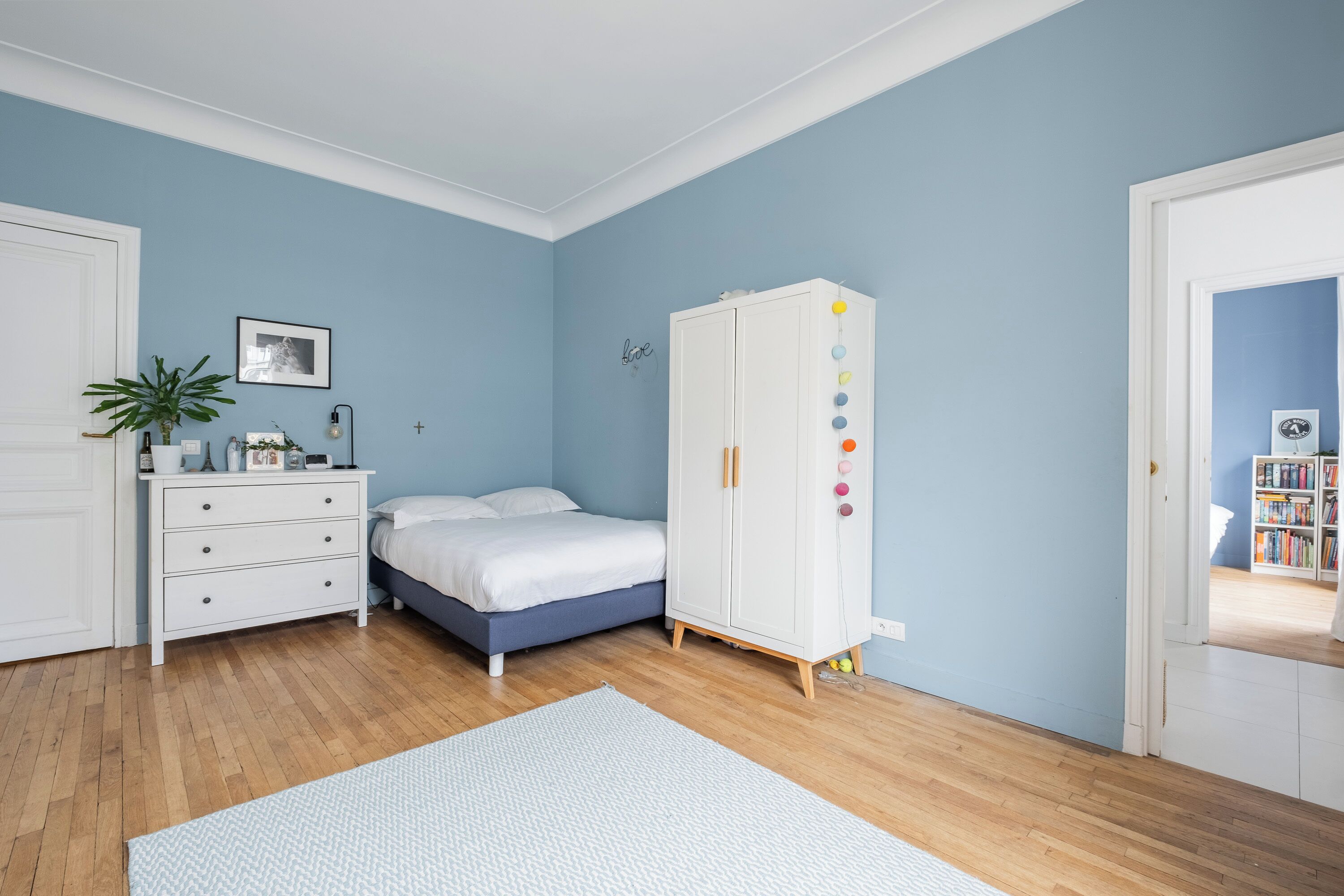 A bedroom with a bed, a white wardrobe, a dresser, a plant, and a bookshelf in the background.