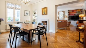 A spacious living room with a wooden floor, a dining area with a large table and chairs, a fireplace, and a bookshelf filled with books. - Triumphant Design, by Veeve (Paris)