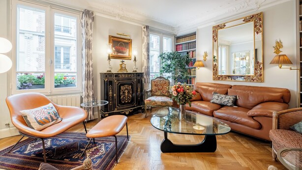 A spacious living room with a leather sofa, a glass coffee table, and a large mirror. There is a bookshelf filled with books and a potted plant. - Triumphant Design, by Veeve (Paris)