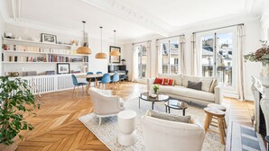 A spacious living room with a sofa, armchairs, a coffee table, and a bookshelf. There is a dining area with a table and chairs, and a large window providing a view of the city. - A Vision in White, by Veeve (Paris)
