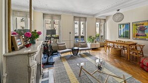 A spacious living room with a fireplace, large windows, a dining area, and a view of the city. - Batignolles Gem, by Veeve (Paris)