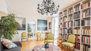 A living room with a sofa, chairs, a round table with a book, a bookshelf, and a large plant. - Literary Radiance, by Veeve (Paris)