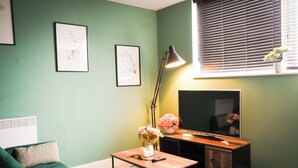 A room with a green wall, a green sofa, a wooden table with a vase of flowers, a floor lamp, and a flat-screen TV. - 40percent off Monthly /2 Bedroom apt / City Centre (Swindon)