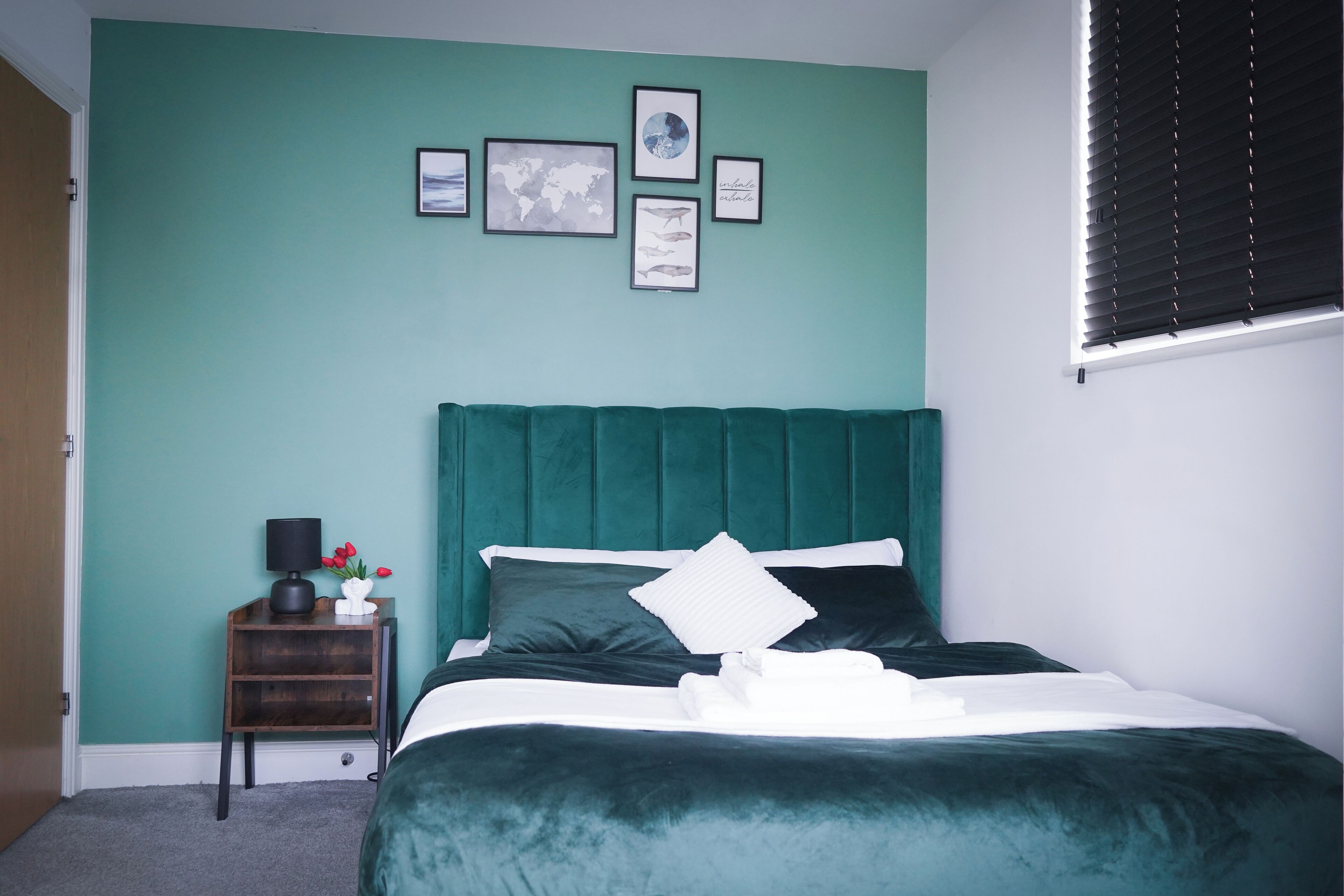 Een slaapkamer met teal wanden, een groen gestoffeerd hoofdeinde, een houten nachtkastje met een lamp en bloemen, en een net opgemaakt bed met witte lakens.