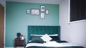 A bedroom with teal walls, a green upholstered headboard, a wooden nightstand with a lamp and flowers, and a neatly made bed with white linens. - 40percent off Monthly /2 Bedroom apt / City Centre (Swindon)