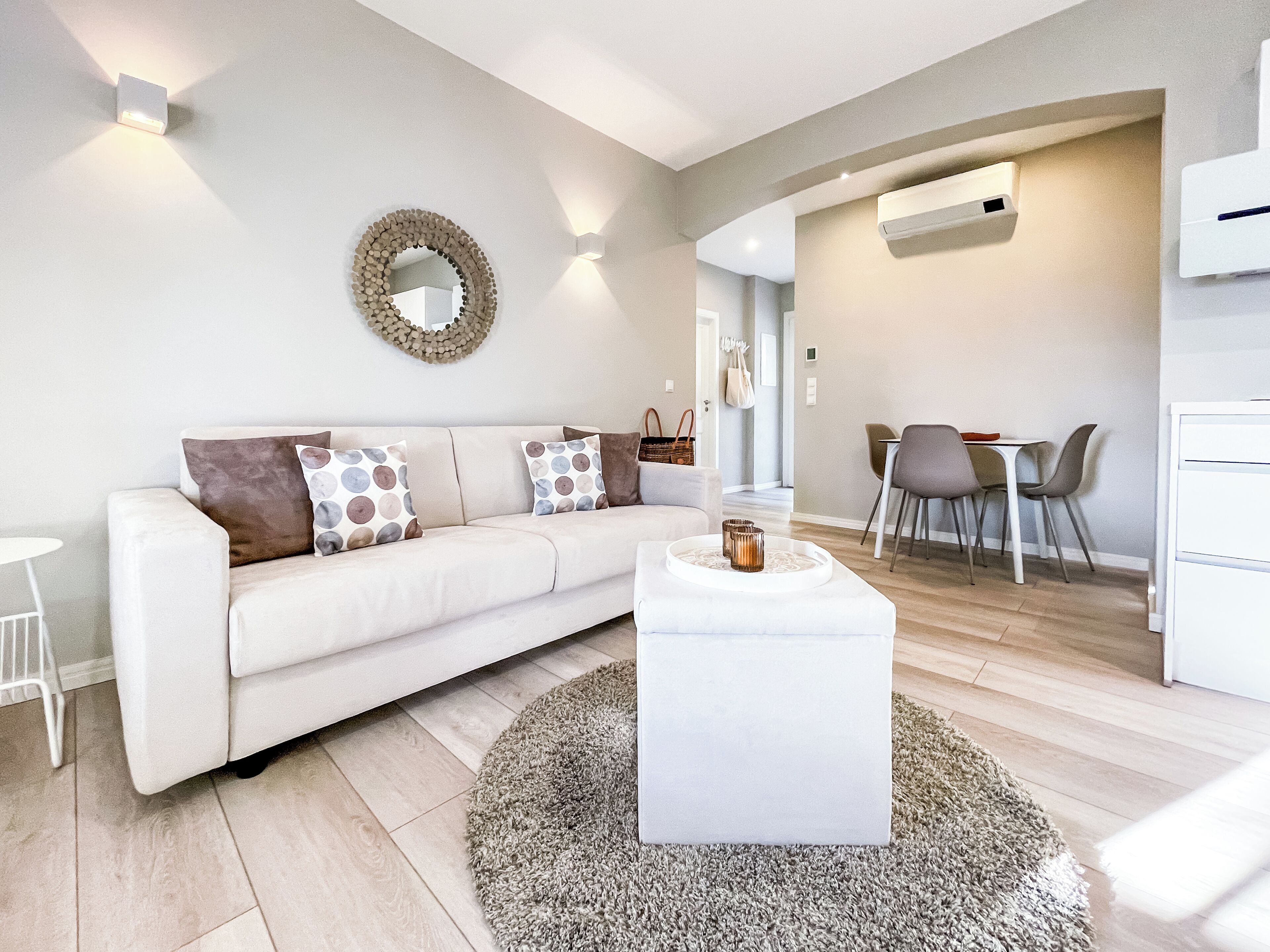 A modern living room with a white sofa, patterned cushions, a round mirror, and a dining area with chairs and a table.