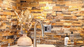 Una cocina con armarios de madera, protector contra salpicaduras de piedra, un lavamanos de acero inoxidable y un florero con flores secas.