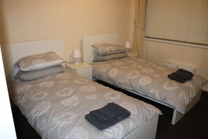 A hotel room with two single beds, each with a nightstand and a lamp. The room has a window with curtains.