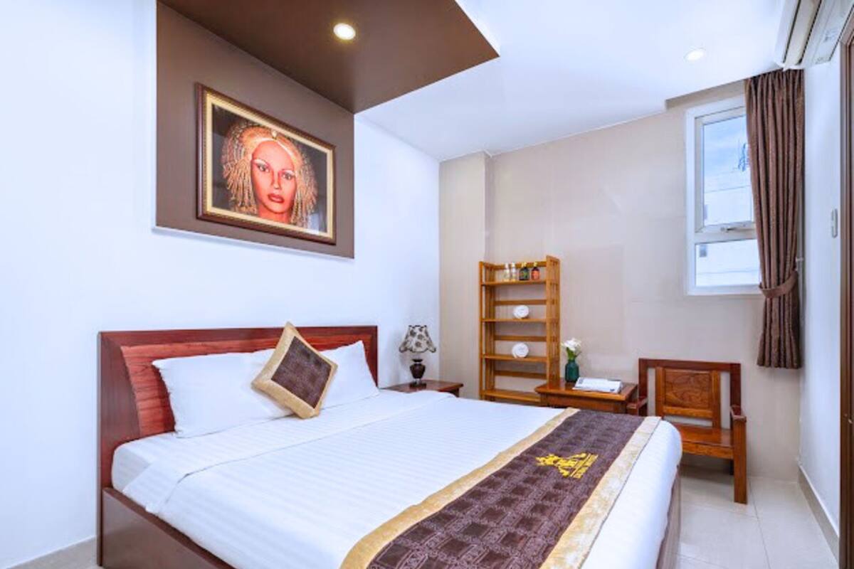 A hotel room with a large bed, a wooden headboard, a bedside table with a lamp, a small shelf with decorative items, and a window with curtains.