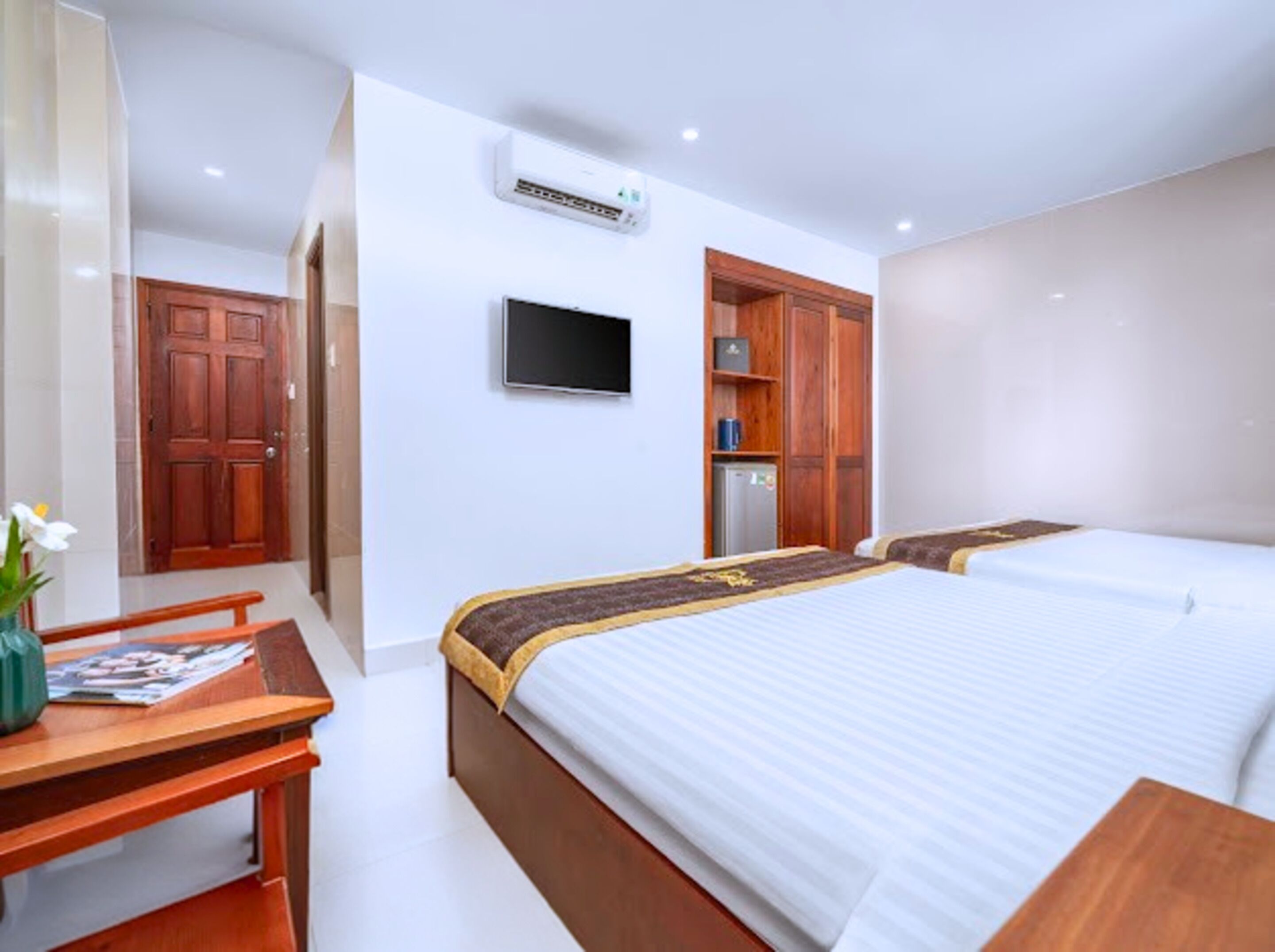 A hotel room with two beds, a wooden desk, a TV mounted on the wall, and a refrigerator.