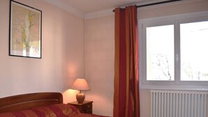 Une chambre à coucher avec un lit, une table de chevet, une lampe, une fenêtre avec des rideaux et un radiateur.
