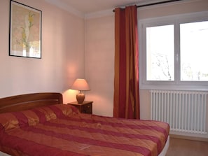 Ein Schlafzimmer mit Bett, Nachttisch, Lampe, einem Fenster mit Vorhängen und einer Heizung.