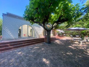 Un edificio con fachada texturizada y amplios ventanales de arco, un árbol en primer plano y una zona de patio empedrada con mesas y sillas al aire libre.