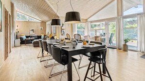Een ruime eethoek met een lange tafel en stoelen, een keuken op de achtergrond en grote ramen met uitzicht op de omgeving.