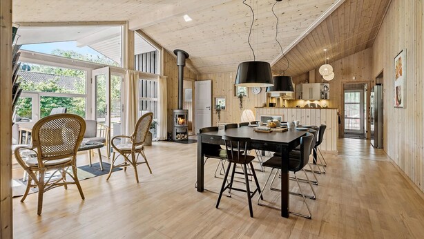 Een ruime kamer met een houten plafond, een centrale eettafel met stoelen, een open haard en grote ramen met uitzicht op de omgeving.