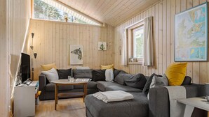 Een knus woongedeelte met een hoekbank, een klein bijzettafeltje en een flatscreen televisie. Er is een raam met gordijnen, een kaart aan de muur en een houten plafond.