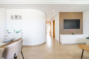 A modern living room with a flat-screen TV mounted on a wall with vertical wood slats, a dining area with a glass table and chairs, and a hallway leading to another room. - Stunning apartment at La Torre Golf Resort MO6022L (La Torre Golf Resort)