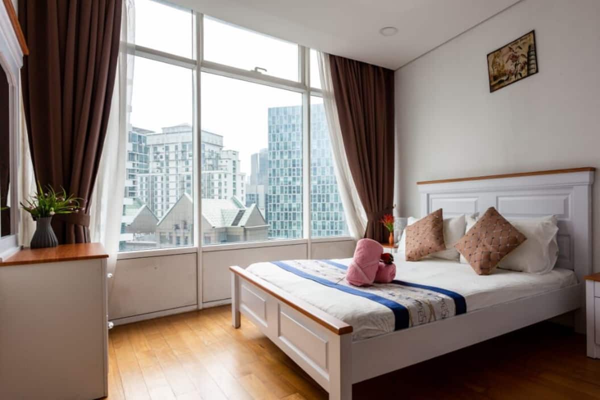 A bedroom with a large window, a bed, a bedside table, and a view of buildings.