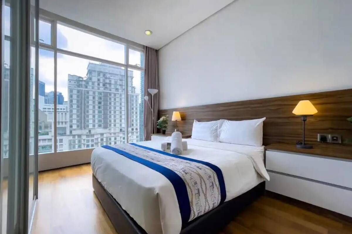 A modern hotel room with a large bed, two bedside lamps, a wooden headboard, and a view of the city through large windows.