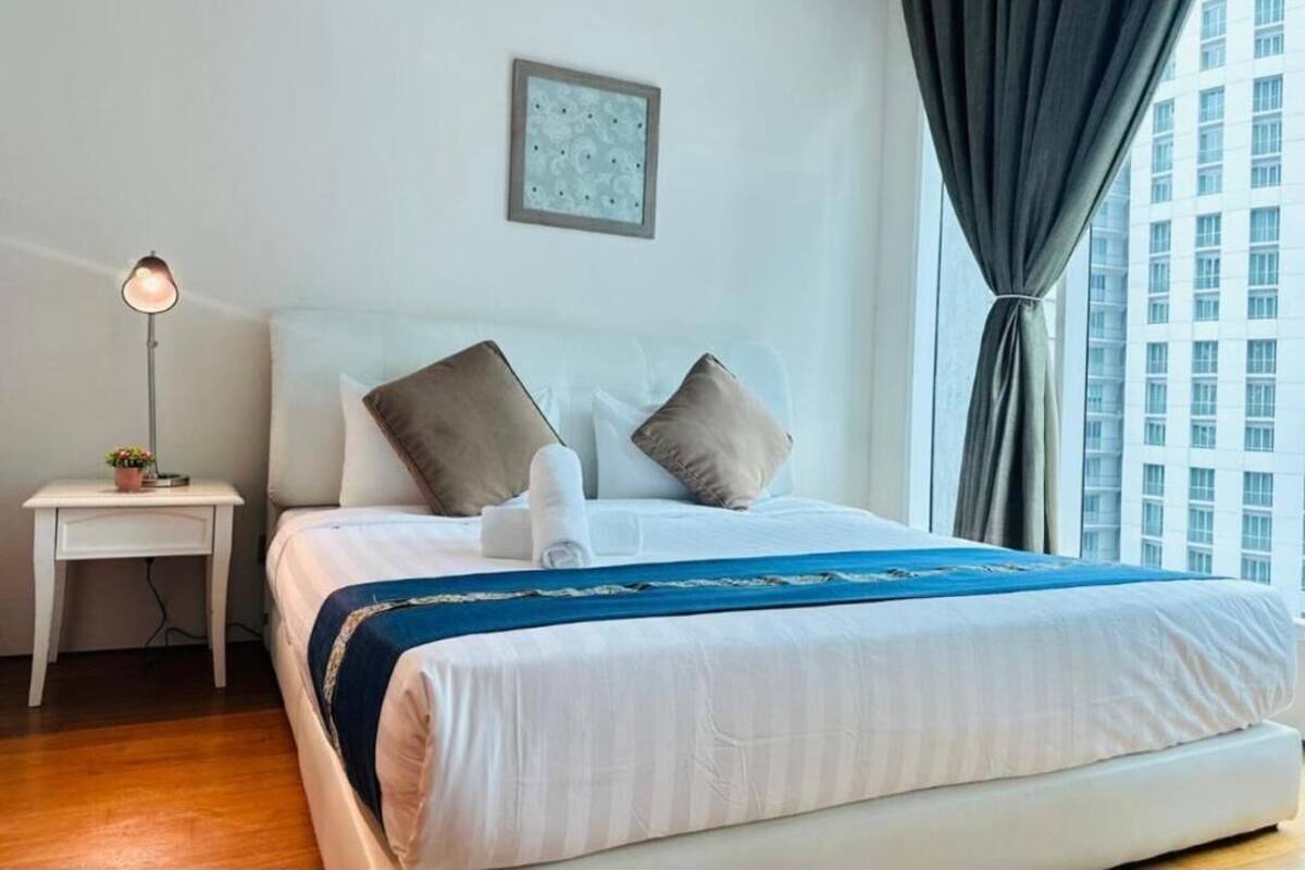 A hotel room with a bed, a bedside table with a lamp, a window with curtains, and a city view.