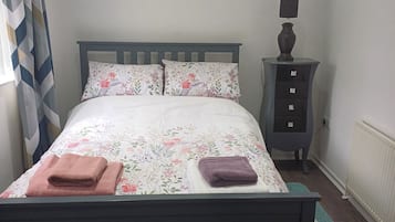 Una cama bien tendida con ropa de cama floral, dos toallas, una mesita de noche con lámpara y un radiador.