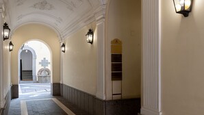 Un pasillo con piso de baldosas, luminarias fijadas en la pared y una puerta con un cartel.