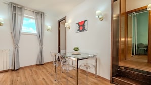 Una habitación con una mesa de comedor de vidrio, sillas traslúcidas, suelo de madera, ventana con cortinas, una lámpara de pared y un espejo.