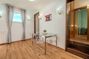 A room with a glass dining table, clear chairs, a wooden floor, a window with curtains, a wall-mounted lamp, and a mirror.