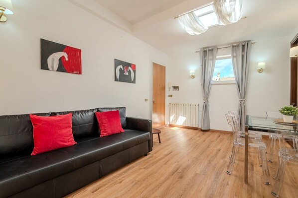 A living room with a black leather sofa, red cushions, a glass dining table with chairs, and two wall-mounted paintings.