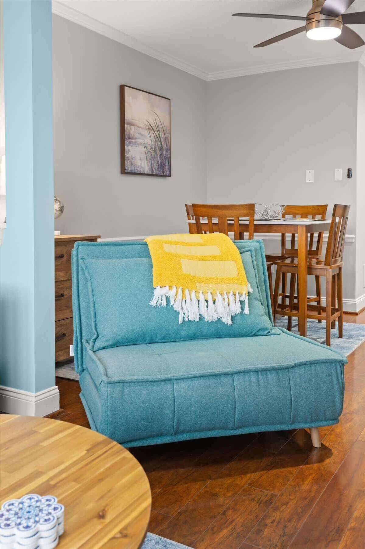 A teal sofa with a yellow throw blanket, a dining table with chairs, and a ceiling fan in a room with wooden flooring and light gray walls.