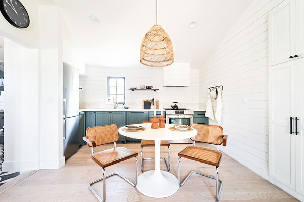 Una cucina moderna con mobili bianchi, un tavolo da pranzo rotondo con quattro sedie in vimini, un lampadario in vimini appeso al soffitto e un orologio sulla parete.