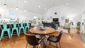 Amplia sala de estar con una mesa redonda de madera, sillas color turquesa y un sofá oscuro. Se observa una zona de cocina con armarios blancos y un refrigerador.