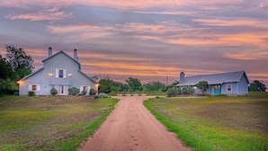 Un camino rural que lleva a una casa de campo, con un sendero de tierra y campos cubiertos de hierba.