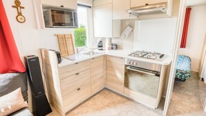 A compact kitchen with a microwave, stove, and sink. There is a small window with blinds, a red curtain, and a bed with a floral bedspread. - Bungalow, terrace, garden, 200m from the beach, by Groom*. (Guidel)