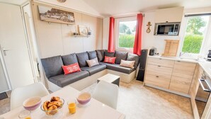 A modern living room with a sectional sofa, a dining table with breakfast items, and a kitchen area with wooden cabinets and a window. - Bungalow, terrace, garden, 200m from the beach, by Groom*. (Guidel)