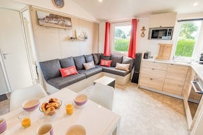 A modern living room with a sectional sofa, a dining table with breakfast items, and a kitchen area with wooden cabinets and a window.