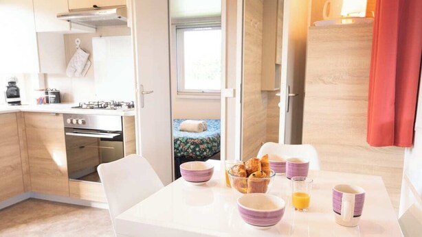 A modern kitchen with white cabinets, a gas stove, and a dining area with a white table set for breakfast. - Bungalow, terrace, garden, 200m from the beach, by Groom*. (Guidel)