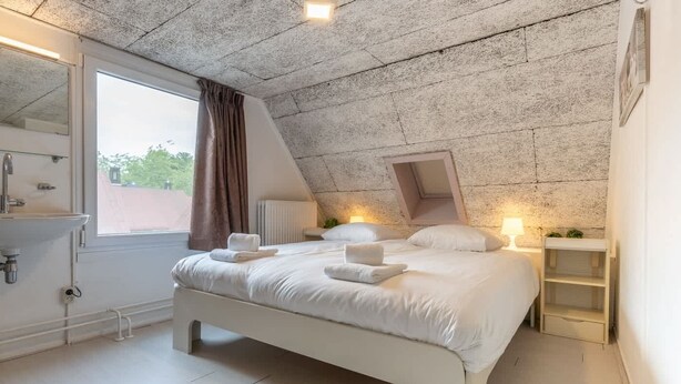 A bedroom with a sloped ceiling, a large bed with white linens, a window with curtains, a radiator, and bedside lamps. - Apartment with balcony in Veere | Buiten de Veste 36 (Veere)
