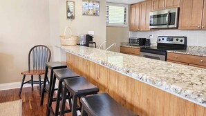 A kitchen with wooden cabinets, a granite countertop, a microwave, a stove, and a bar area with stools. - Litchfield by the Sea - 3 BR Villa - View of Heron Marsh Lake (Pawleys Island)