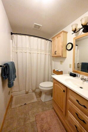 A bathroom with a toilet, a sink, wooden cabinets, a mirror, a clock, and a shower curtain. - Cabin of Hygge: 19 Miles to Yellowstone (Island Park)