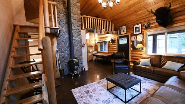 A log cabin interior with a wooden staircase, a stone wall, a fireplace, a dining area, and a living room with a sofa and a bench. - Cabin of Hygge: 19 Miles to Yellowstone (Island Park)