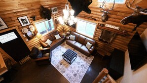 A log cabin interior with a wooden floor, a leather sofa, a coffee table, a fireplace, and mounted animal heads. - Cabin of Hygge: 19 Miles to Yellowstone (Island Park)
