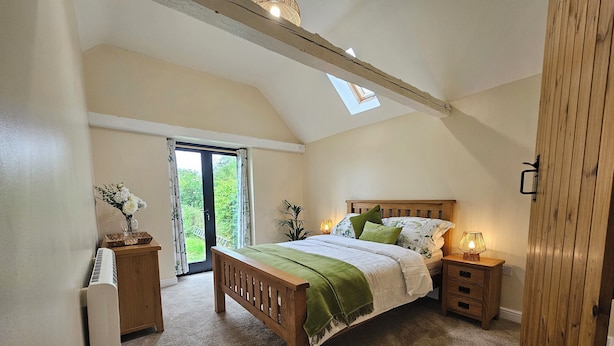 A bedroom with a sloped ceiling, a bed with a green bedspread, two bedside tables with lamps, a wooden nightstand, a window with curtains, and a view of greenery outside. - Lily's Cottage Suffolk Sleeps 2 With Pets (Stowmarket)