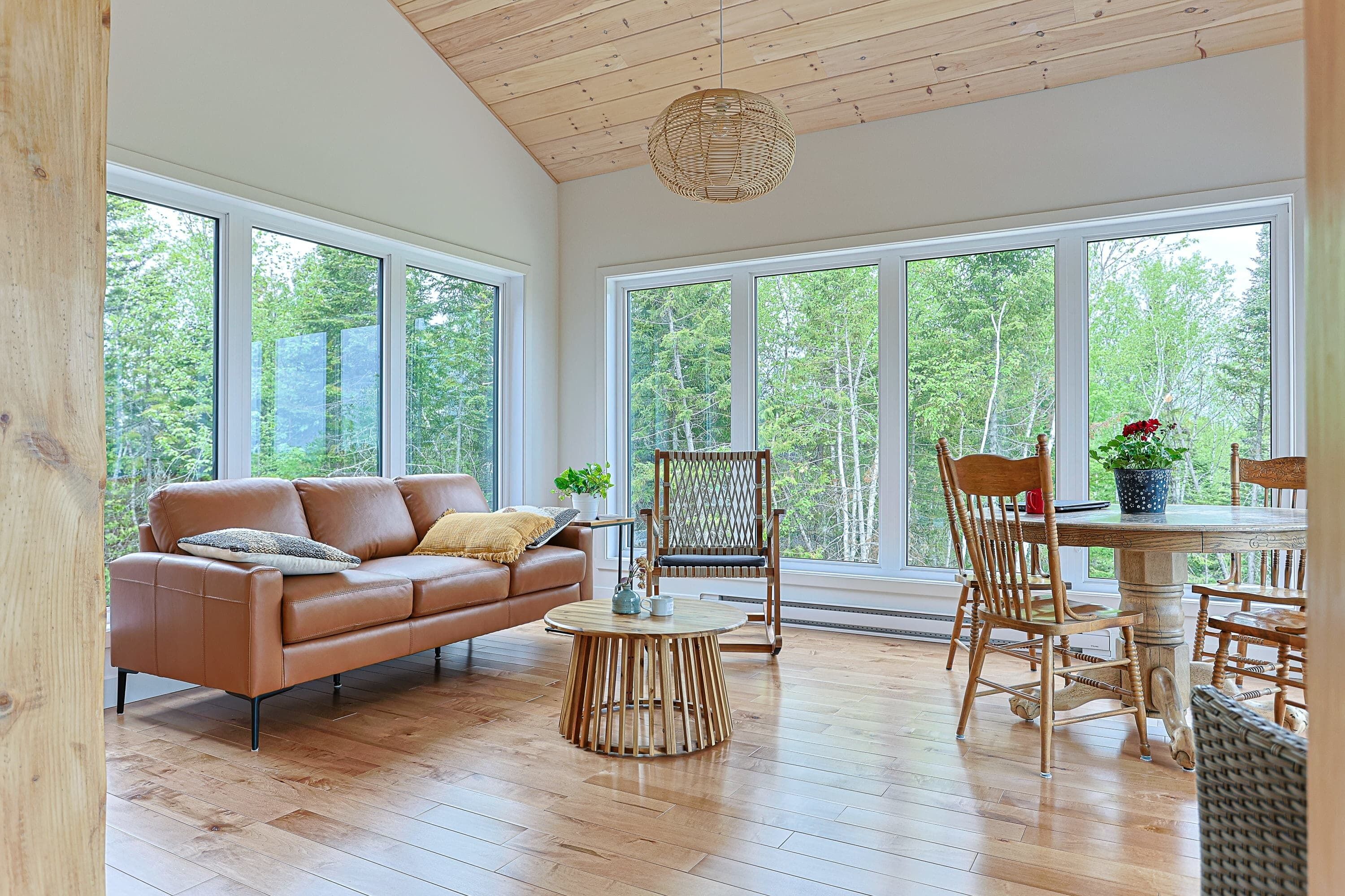 A spacious room with a wooden floor, a brown leather sofa, a round wooden coffee table, and a dining table with chairs. Large windows provide a view of the outdoors.