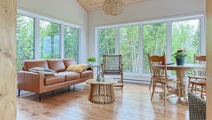 Ruime kamer met houten vloer, een bruine leren bank, een ronde houten salontafel en een eettafel met stoelen. Grote ramen bieden uitzicht op het omliggende landschap.