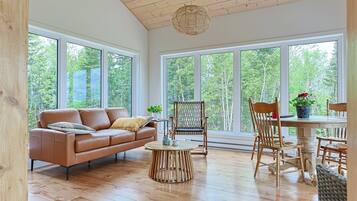 A spacious room with a wooden floor, a brown leather sofa, a round wooden coffee table, and a dining table with chairs. Large windows provide a view of the outdoors.